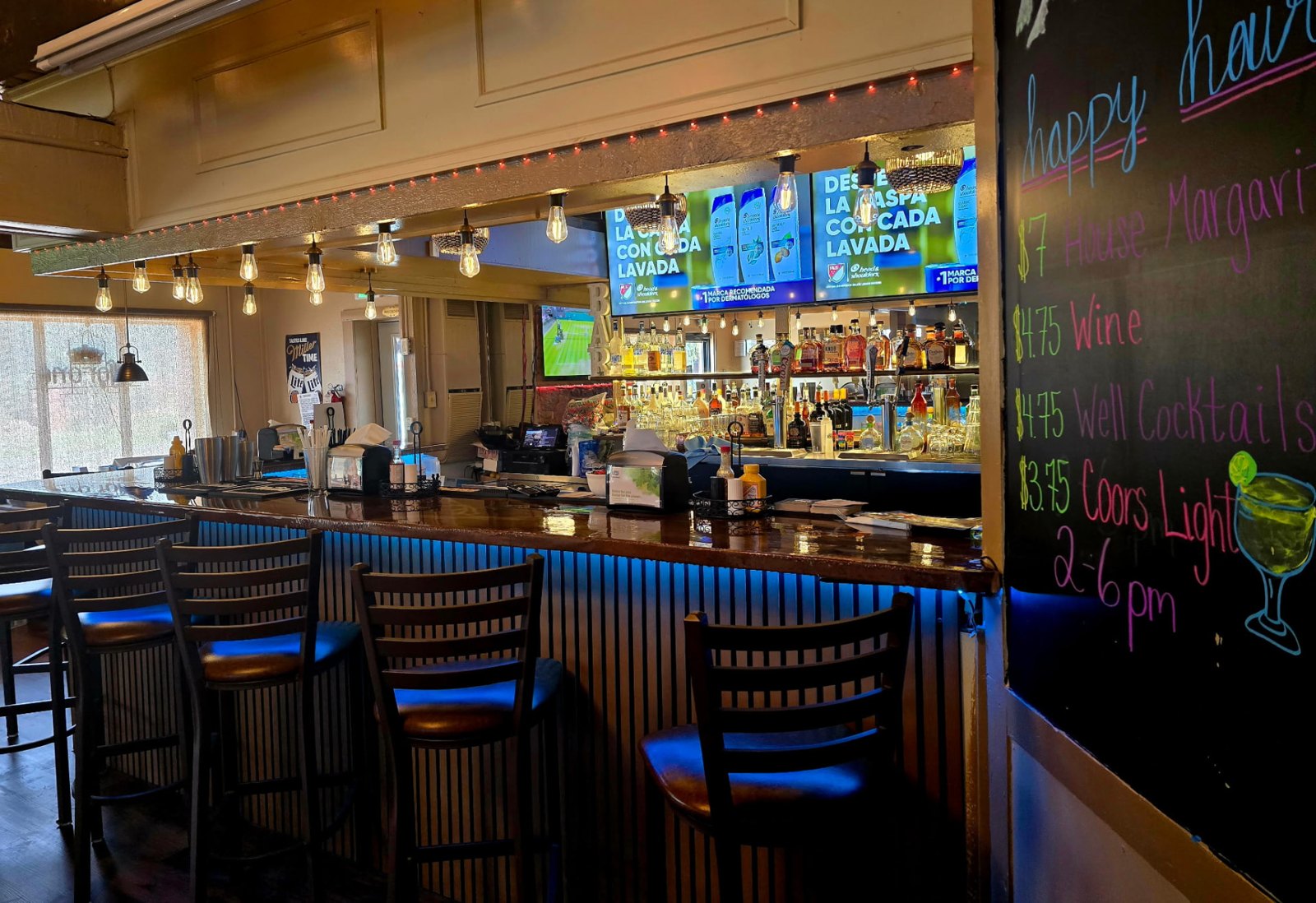 String lights over the Vista Lounge bar with a hand-lettered happy hour chalkboard listing margaritas, wine and Coors Light