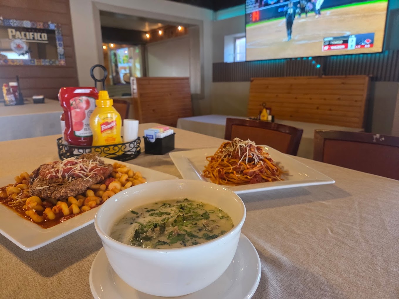 Chicken parmesan over pasta, spaghetti with meatballs and a bowl of creamy Toscana soup on a table with the game on TV