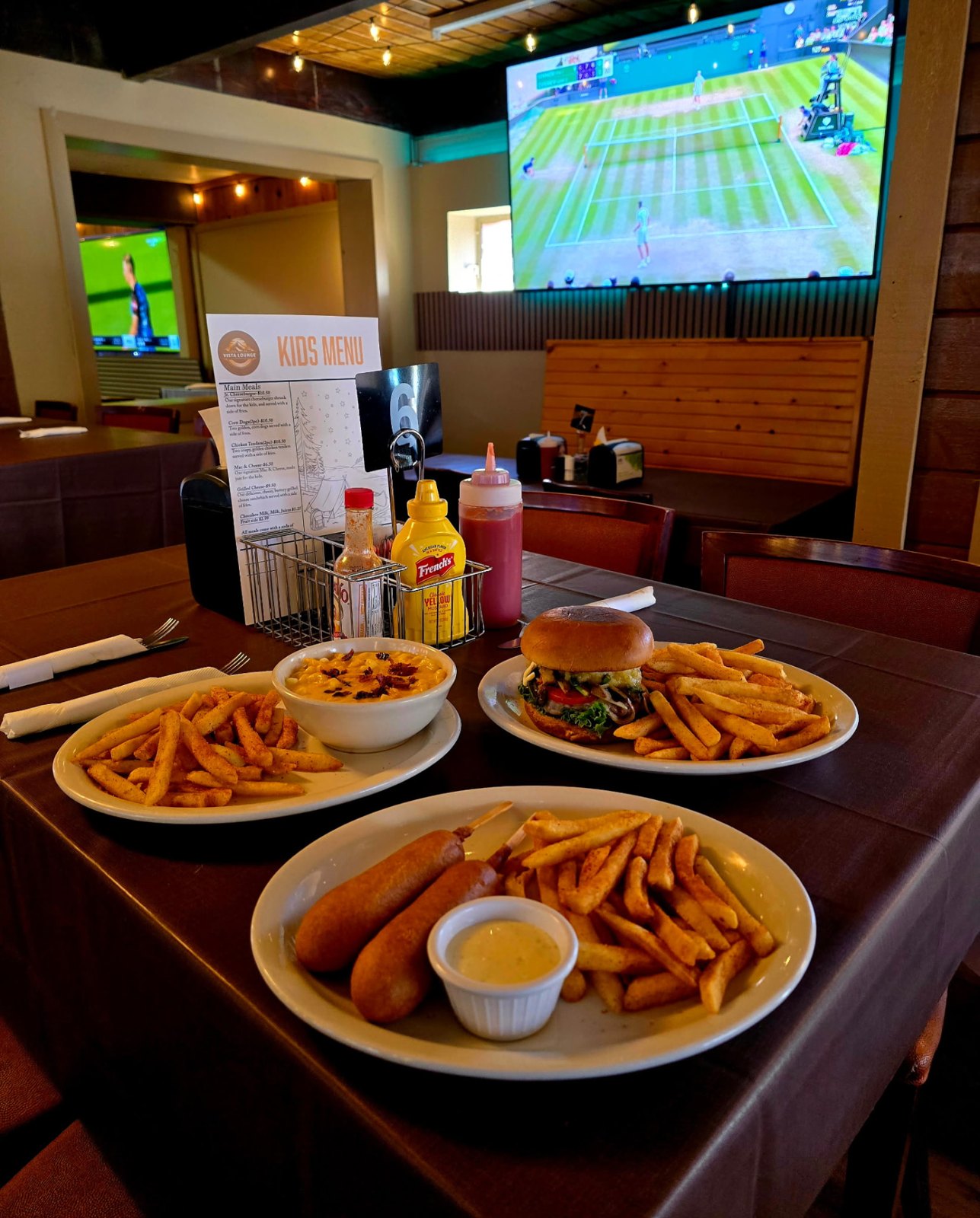 Burger, fries, loaded mac and cheese and corn dogs on a dining table with the match on the big screen behind